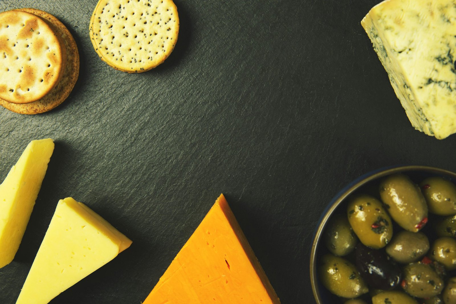A delectable cheese platter featuring various cheeses, crackers, and olives on a slate background.